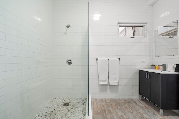 A bathroom with white tiled walls, a glass shower enclosure, a wooden vanity with a mirror, and a window.