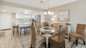 A modern kitchen with white cabinets, a central island, and a dining area with wicker chairs and a glass table. - Oceanfront 1st-Floor Oasis: Steps to Pool & Beach (Fernandina Beach)