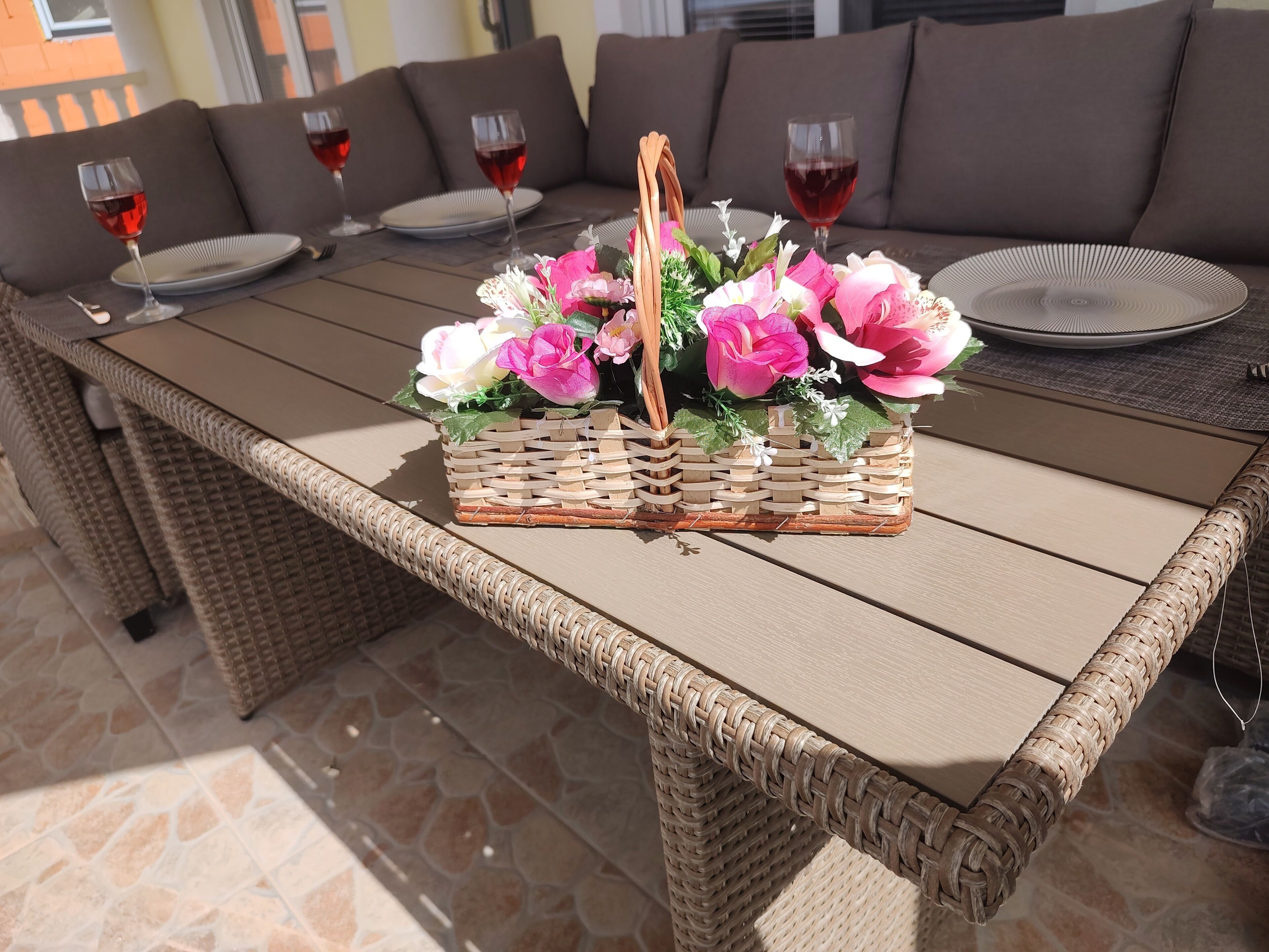 A wicker table with a woven basket containing pink and white flowers, set on a patio with a stone floor.