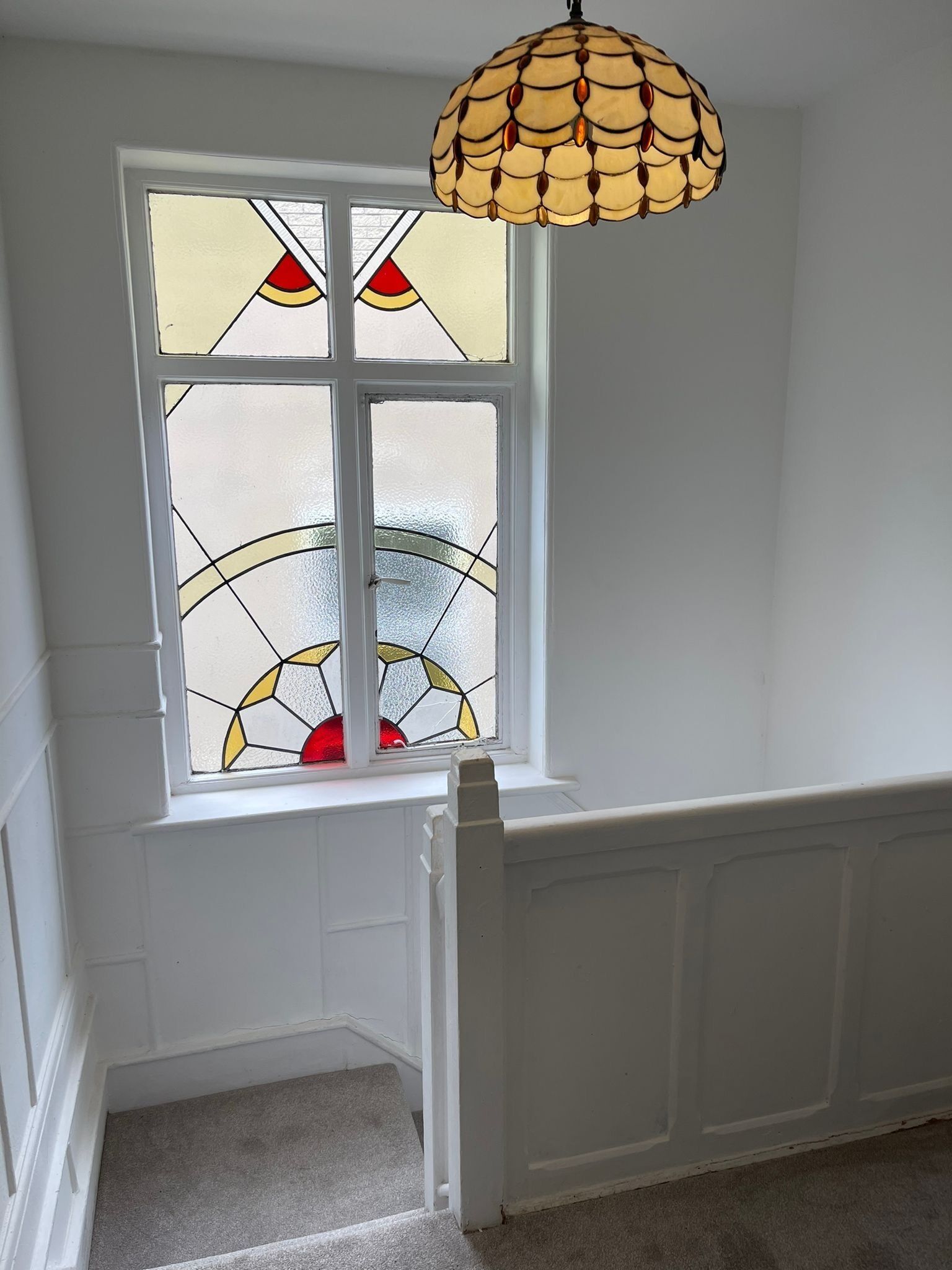 A corner room with a white paneled wall, a window with stained glass, and a hanging light fixture.