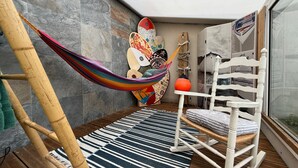 Una hamaca en un rincón, una mecedora blanca, un tapete a rayas y una tabla de surf contra la pared.