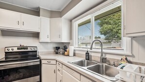 A modern kitchen with stainless steel appliances, white cabinetry, a double sink, and a window with blinds. - NEW-Parrot Head Haven-3BR-1BA-Themed-Bar-Patio (Piqua)