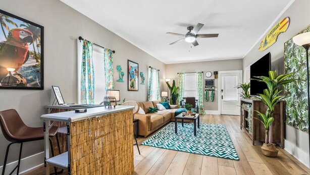 A modern living room with a wooden floor, a sofa, a coffee table, and a dining area with a bamboo partition. There are plants and artwork on the walls. - NEW-Parrot Head Haven-3BR-1BA-Themed-Bar-Patio (Piqua)