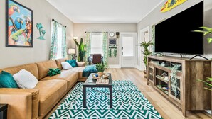 A living room with a brown leather sofa, a patterned rug, a wooden TV stand, and a large painting on the wall. - NEW-Parrot Head Haven-3BR-1BA-Themed-Bar-Patio (Piqua)
