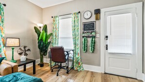 A room with a bed, a desk, a chair, a plant, a clock, and a door with blinds. - NEW-Parrot Head Haven-3BR-1BA-Themed-Bar-Patio (Piqua)