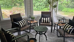 A covered patio with wicker furniture, a round table, and large windows offering a view of greenery.