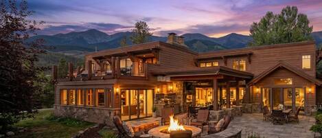 A large, multi-level house with a fire pit and outdoor seating area, surrounded by a landscaped garden and mountains in the background.