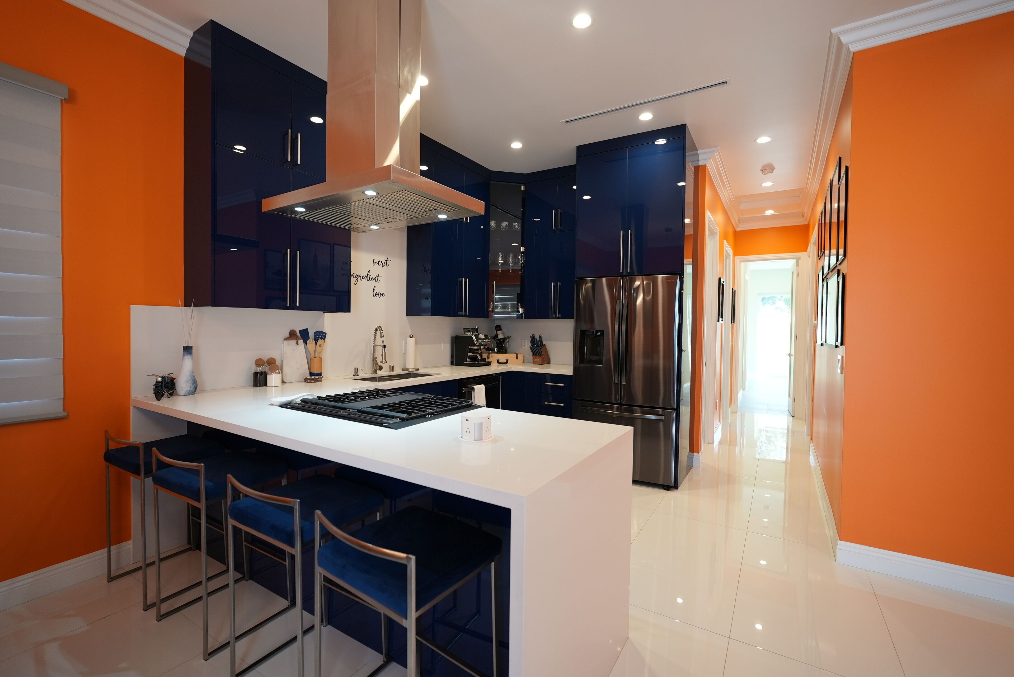 Una cocina moderna con armarios azul oscuro, una isla blanca con taburetes de bar, electrodomésticos de acero inoxidable y una pared de acento anaranjada brillante.