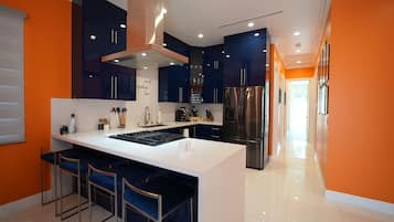 Una cocina moderna con armarios azul oscuro, una isla blanca con taburetes de bar, electrodomésticos de acero inoxidable y una pared de acento anaranjada brillante.