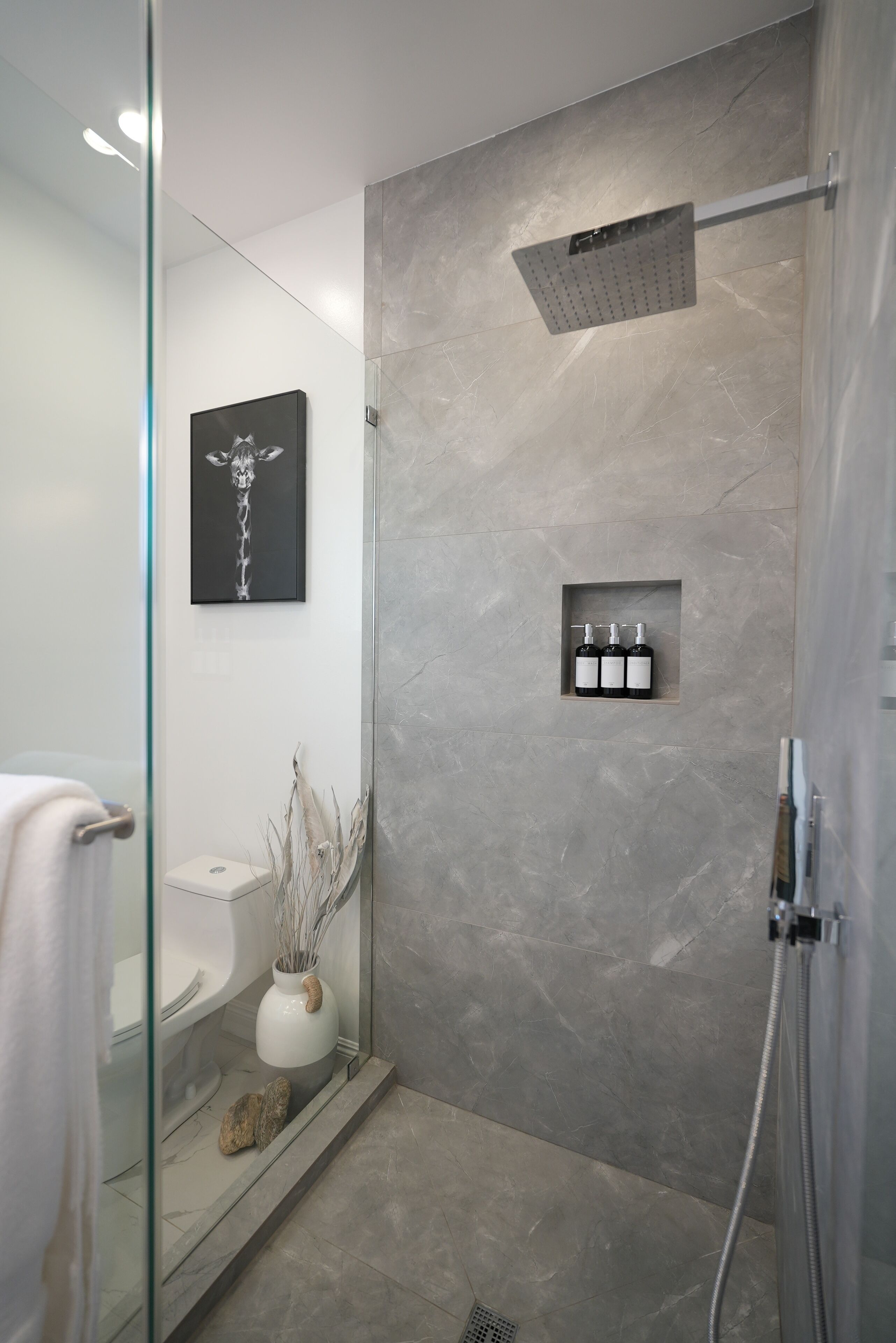 Un baño moderno con cabina de ducha de vidrio, pared de mármol, inodoro, lavamanos, un florero con plantas secas y un cuadro en la pared.