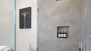 Un baño moderno con cabina de ducha de vidrio, pared de mármol, inodoro, lavamanos, un florero con plantas secas y un cuadro en la pared.