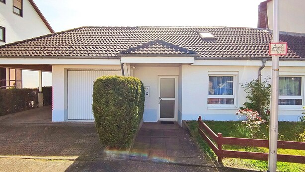 Ein einstöckiges Haus mit Ziegeldach, einer Haustür und einem kleinen Garten mit Hecke und Blumen.