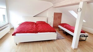 Ein Schlafzimmer mit einem großen Bett, einem kleineren Bett, einem Heizkörper, einem Fenster und einer Uhr an der Wand.