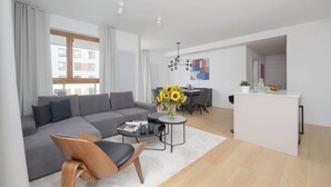 A modern living room with a grey sofa, a wooden chair, a coffee table, and a dining area with a white table and chairs. - Spacious Apartment | 3 Bedrooms | Parking (Warszawa)