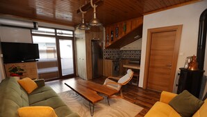 Ein modernes Wohnzimmer mit einer Couch, Sesseln und einem Couchtisch. Es gibt eine Küchenzeile mit Herd, Spüle und Schränken. Ein Fernseher ist an der Wand montiert, und eine Schiebetür führt in einen weiteren Raum.