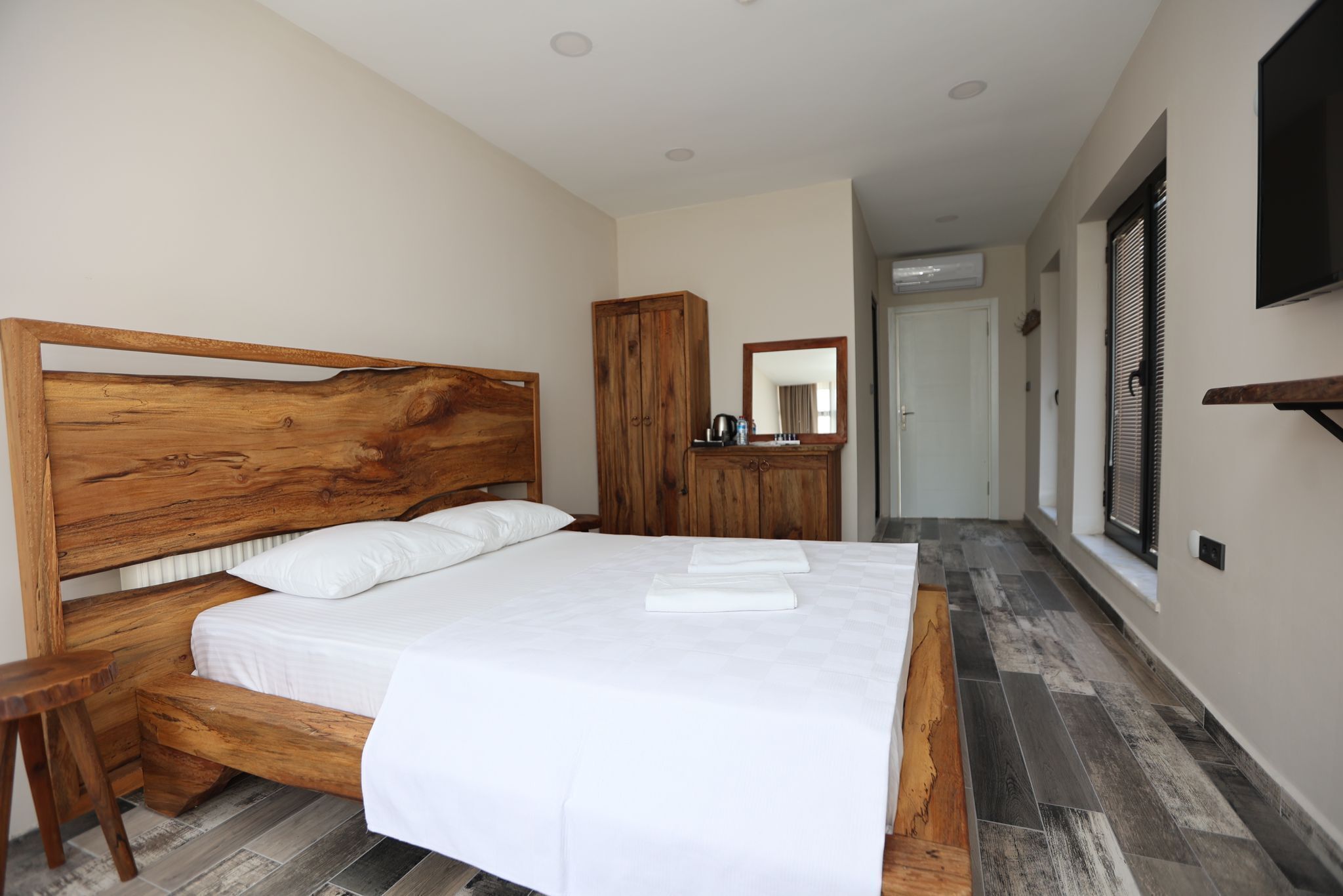 Un dormitorio con cabecera de madera, cama con ropa de cama blanca, armario de madera, espejo y televisión en la pared.