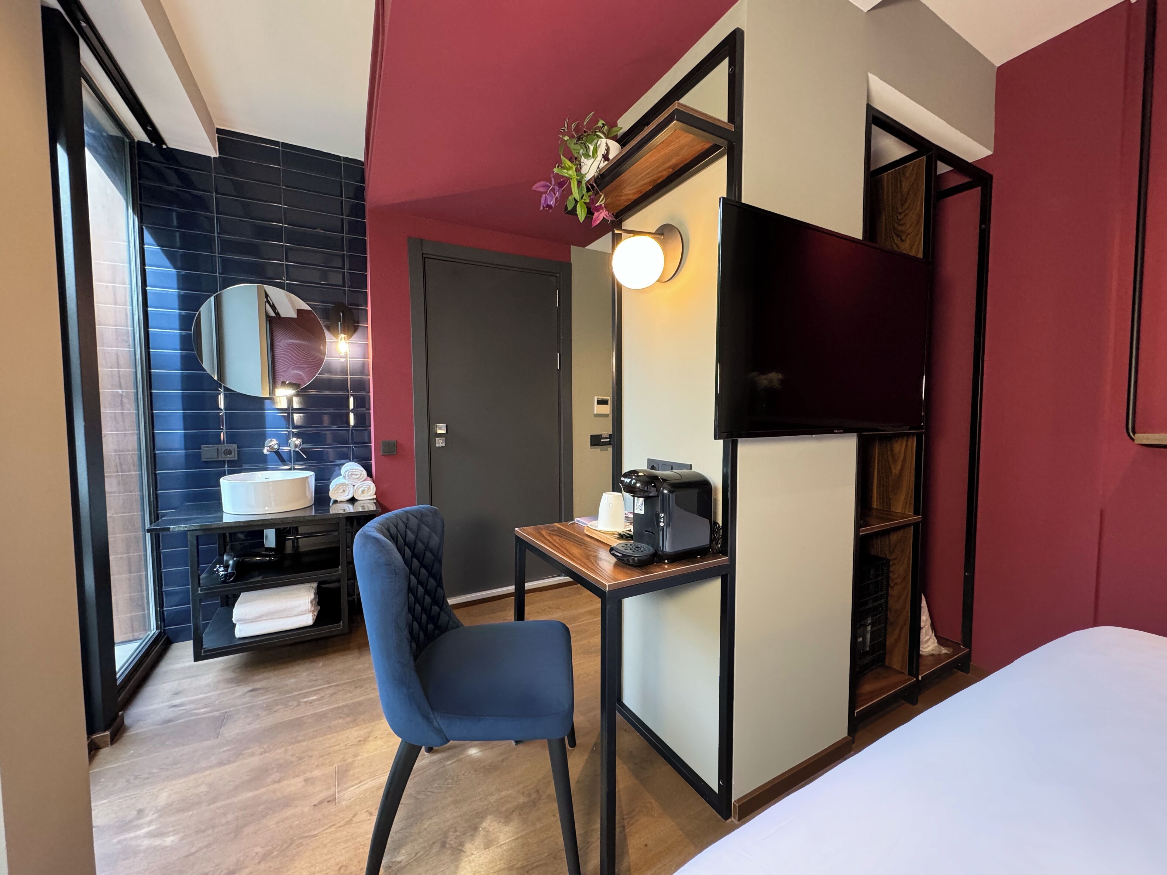 A hotel room with a bed, a desk with a coffee maker, a chair, a TV, a mirror, and a bathroom with a sink and towel rack.
