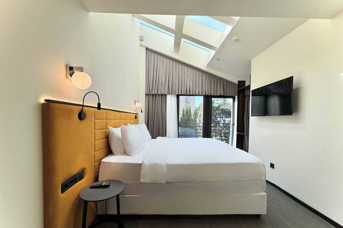 A modern hotel room with a large bed, a bedside table, a TV mounted on the wall, and a skylight above the bed.