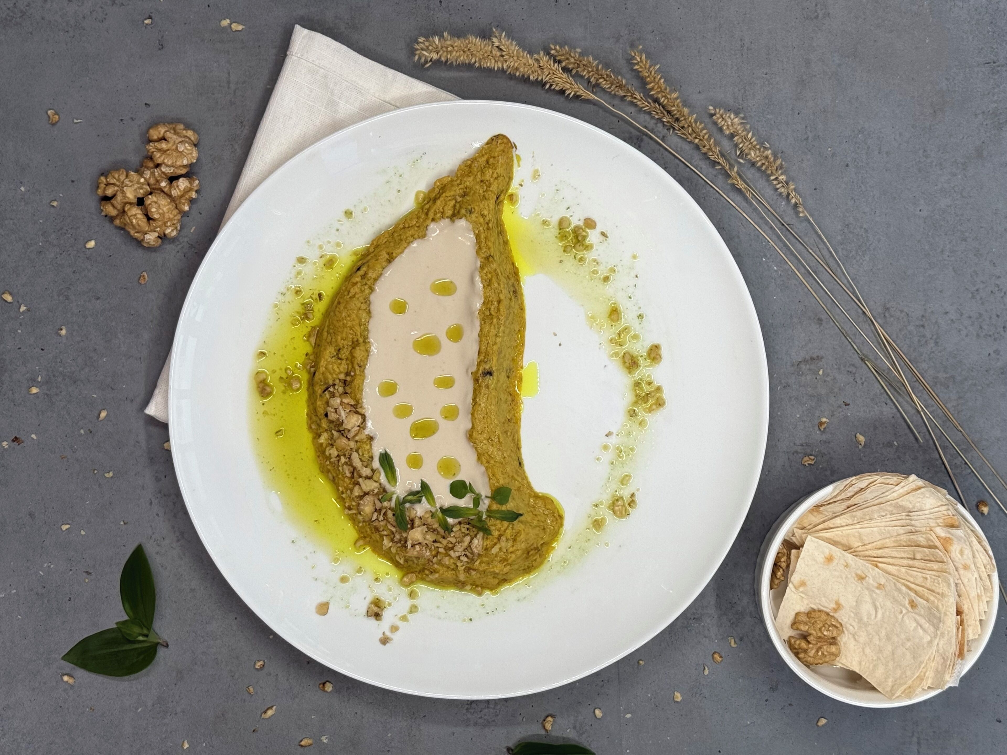 Un piatto con un cibo, una ciotola di noci e alcune foglie su una superficie grigia.
