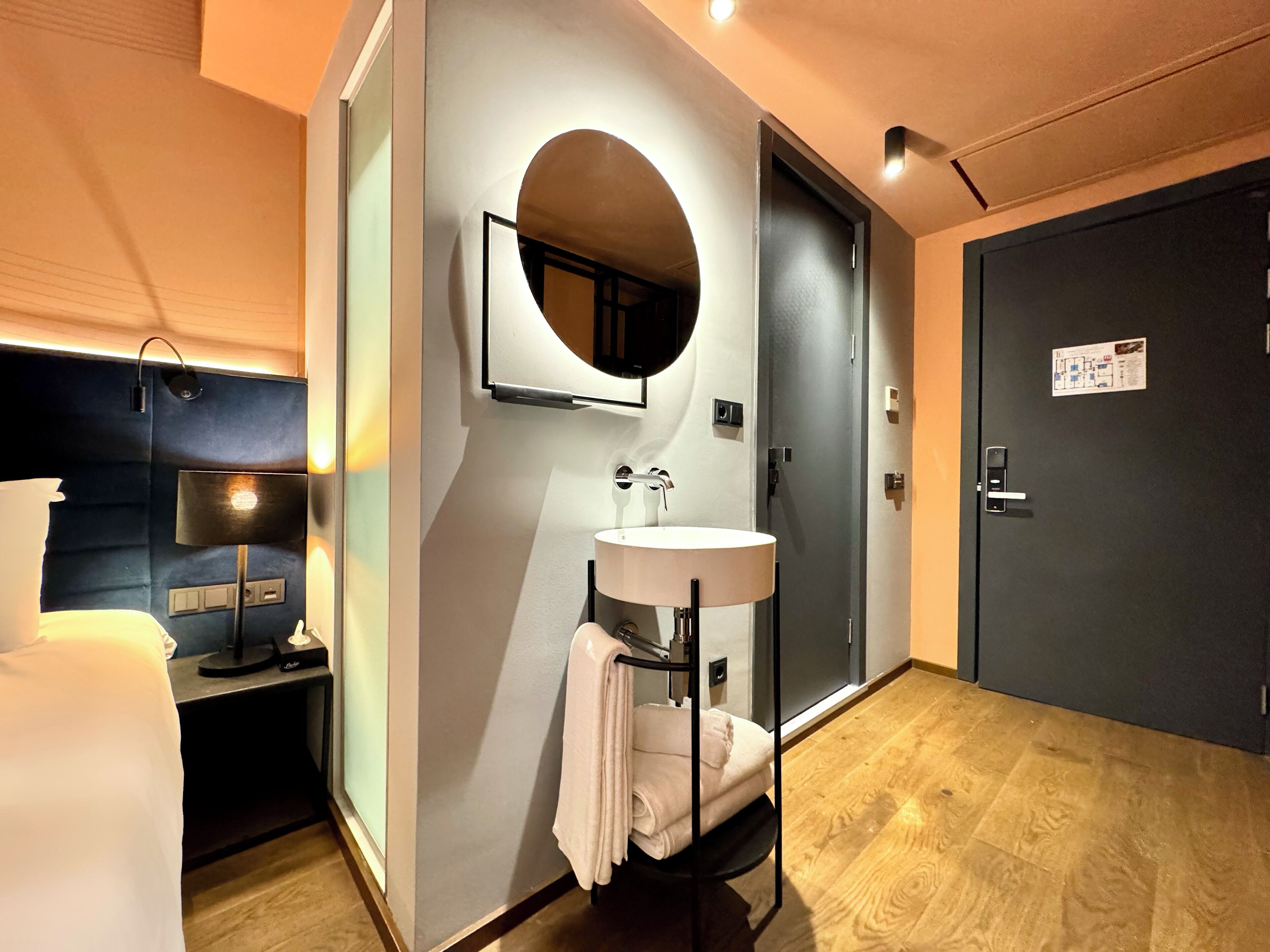 A hotel room with a bed, bedside table, lamp, wall-mounted mirror, sink, towel rack, and a door with a map sign.