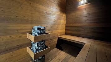 A sauna room with wooden walls and a bench. There is a wooden shelf with a black container holding towels.