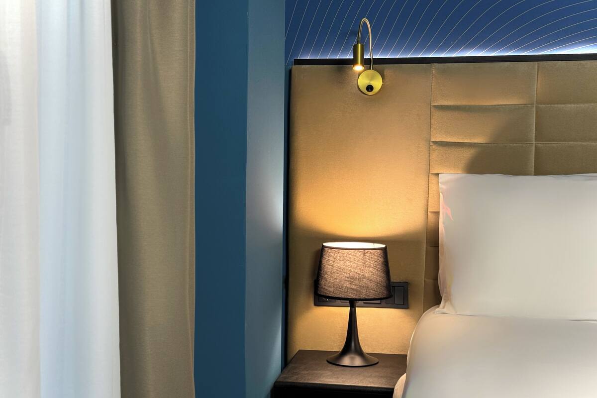 A hotel room with a bed, a nightstand with a lamp, and a wall-mounted light fixture.