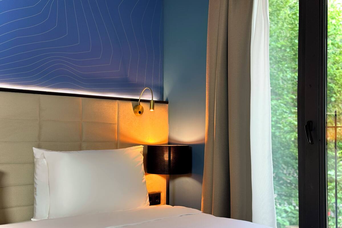 A hotel room with a bed, a nightstand, a lamp, a window with curtains, and a wall with a blue design.