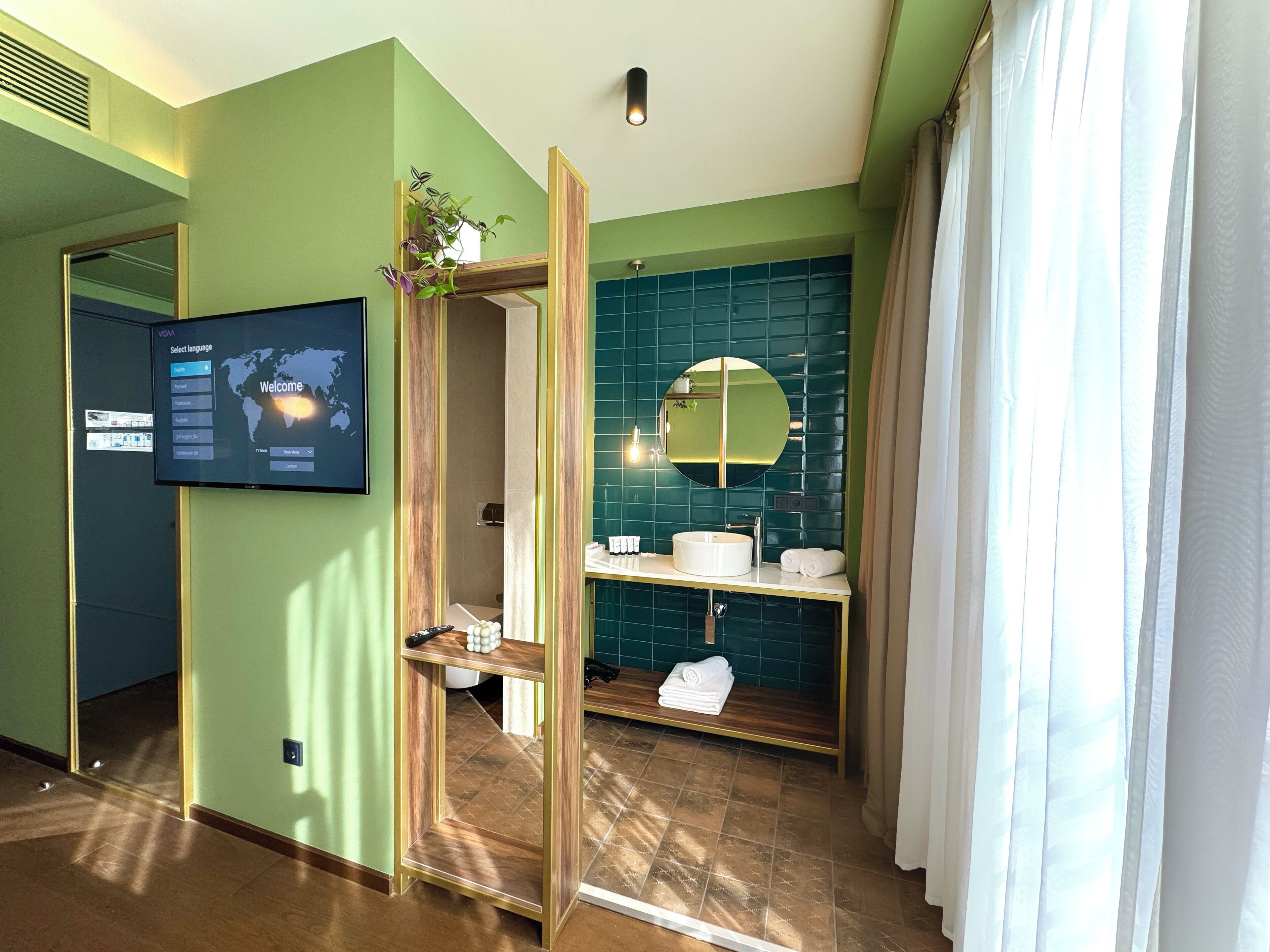 A modern bathroom with a green wall, a large mirror, a sink, and a toilet. There is a TV mounted on the wall displaying a language selection screen.