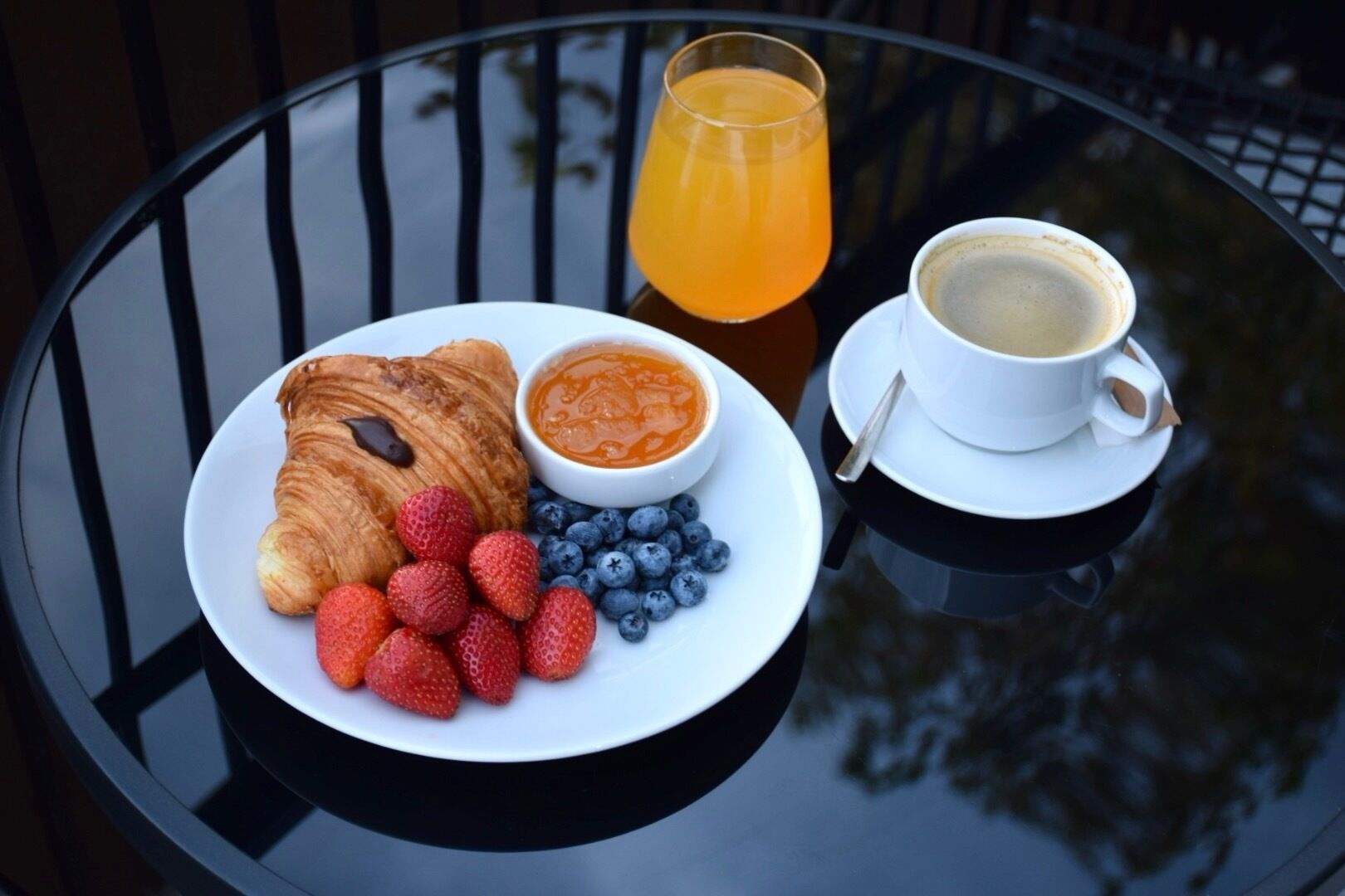 Une table ronde dressée pour le petit-déjeuner, avec un croissant, des fraises, des myrtilles, un bol de confiture, un verre de jus d’orange et une tasse de café.