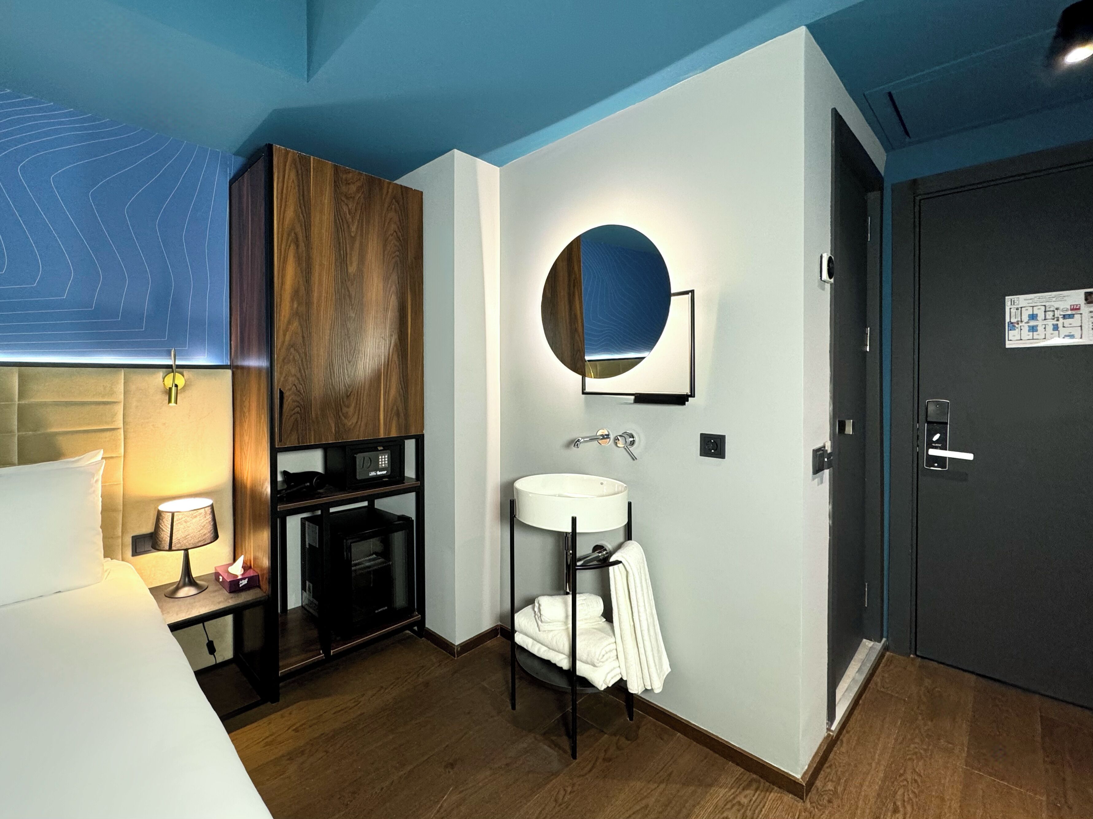 Une chambre d’hôtel moderne avec une tête de lit en bois, un miroir rond, un lavabo blanc et une porte sombre.