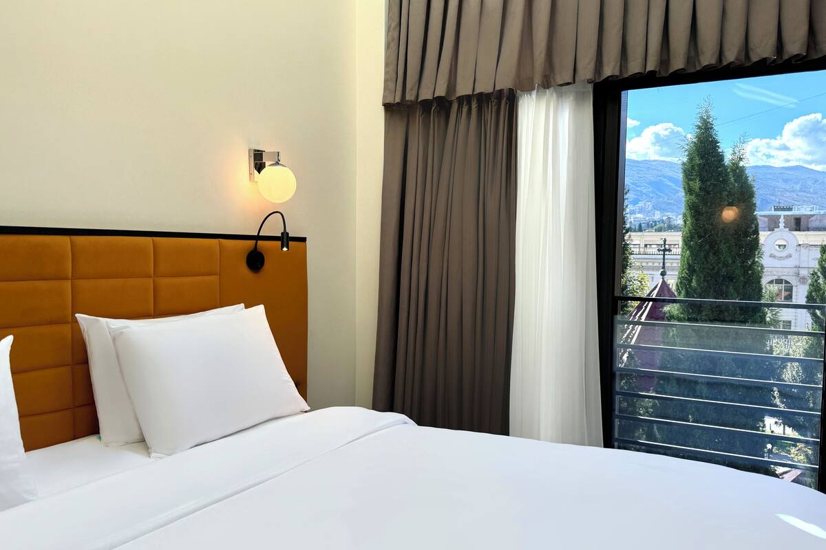 A hotel room with a large bed, a headboard, a wall-mounted light, and a view of a cityscape through a window with curtains.