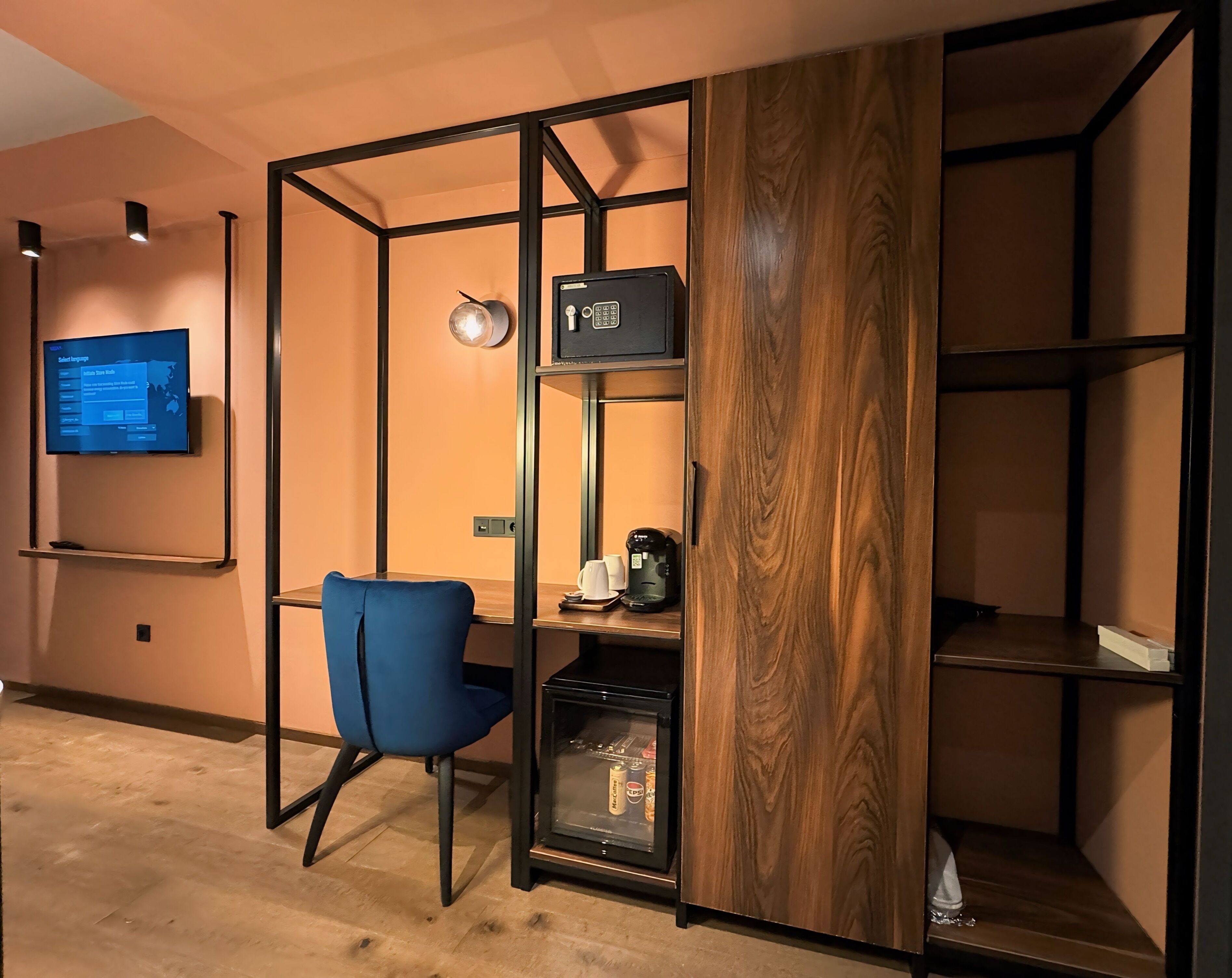 Une chambre moderne avec un fauteuil bleu, un téléviseur fixé au mur, une machine à café et un réfrigérateur.
