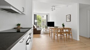 Una cocina moderna con encimera negra, armarios blancos y estufa integrada. Hay una zona de comedor con mesa y sillas de madera, y un televisor montado en la pared.