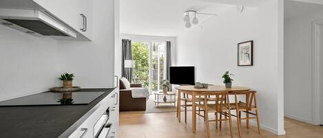 Een moderne keuken met een zwart aanrecht, witte kasten en een inbouwfornuis. Er is een eethoek met een houten tafel en stoelen, en een aan de muur bevestigde televisie.