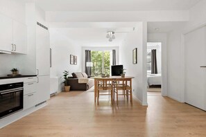 Una cocina moderna con armarios blancos, un comedor con una mesa de madera, y una sala de estar con sofá y televisor.