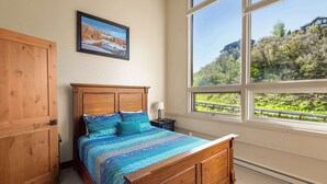 A bedroom with a wooden bed, a nightstand, a lamp, a window with a view of greenery and buildings, and a wooden wardrobe. - Vista Viewhaus Lodge (Steamboat Springs)