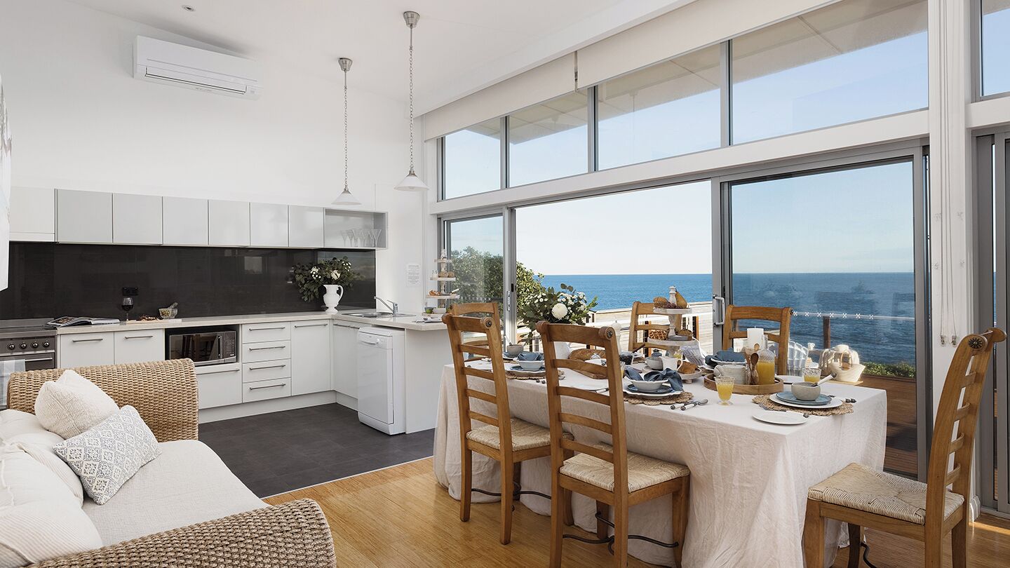 A modern kitchen with white cabinetry, a dining area with wooden chairs, and large windows overlooking the sea.