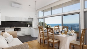 Una cocina moderna con armarios blancos, una zona de comedor con sillas de madera y amplios ventanales con vistas al mar.
