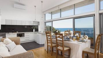 A modern kitchen with white cabinetry, a dining area with wooden chairs, and large windows overlooking the sea.