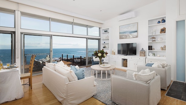 Amplia sala de estar con un gran ventanal que da a un balcón con vistas al mar. Hay un sofá blanco, una mesa redonda de centro y un televisor sobre un mueble blanco.