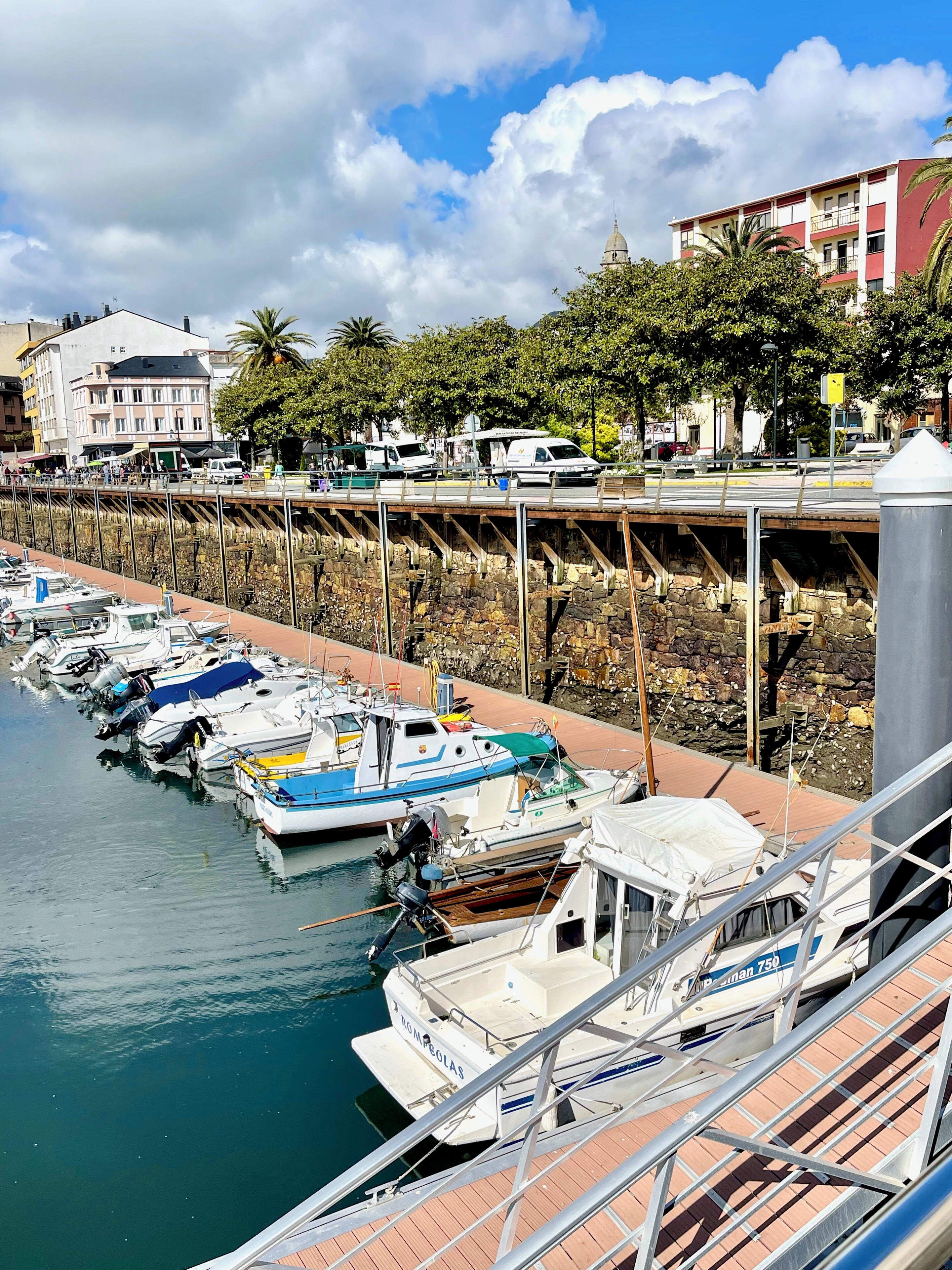 Una marina con barche ormeggiate lungo un molo in pietra, un ponte sopra e edifici sullo sfondo.