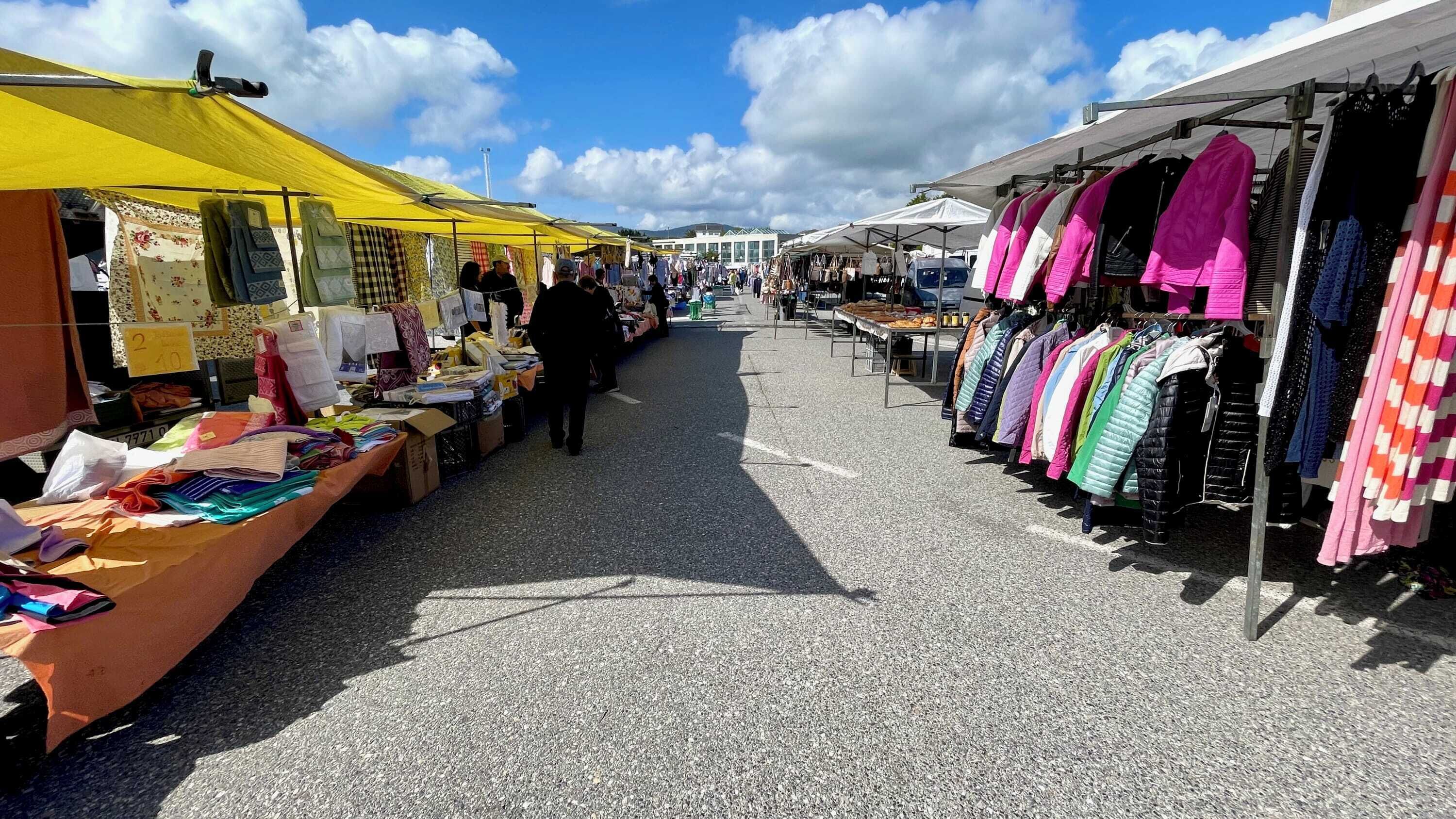 Een markt met kraampjes die textiel en kleding verkopen, onder gele marktkleden.