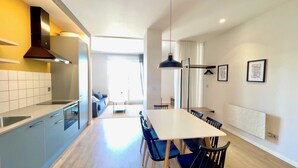 A modern kitchen with a built-in stove, a dining area with a white table and blue chairs, and a large window providing natural light. - The Cliffs  A Ribeira - Areoura (Foz)