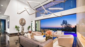 A modern living room with a large sofa, armchairs, and a coffee table. There is a large window with a city view and a sculpture on the wall.