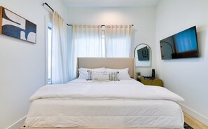 A neatly made bed with white bedding, a bedside table with a lamp, and a wall-mounted TV.