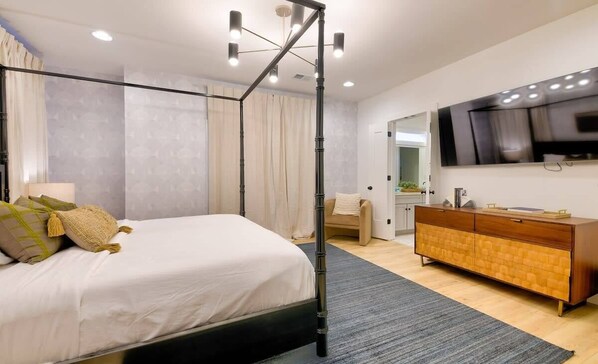 A modern bedroom with a four-poster bed, a wooden dresser, a flat-screen TV, and a large mirror.