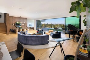 A modern living room with a large window, a round seating area, a piano, and a dining table. - Dolce Vista - Bel Air Estate | 270° Views, Pool & Theater (Bel Air, Los Angeles)