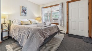 Un dormitorio con cama, mesita de noche, lámpara, ventana con cortinas y puerta.