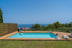 Una piscina rettangolare con un pontile in legno, un'area relax con cuscini e vista sul mare.