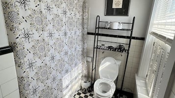Un baño con inodoro, cortina de ducha estampada con motivos florales, suelo a cuadros blanco y negro y un estante con artículos de aseo.
